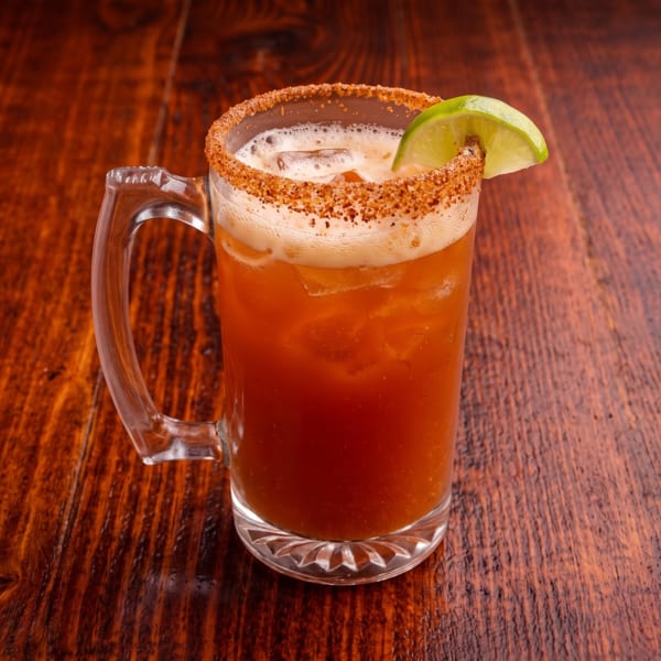 Michelada in a glass mug with a spiced rim and lime wedge on a dark wooden table.