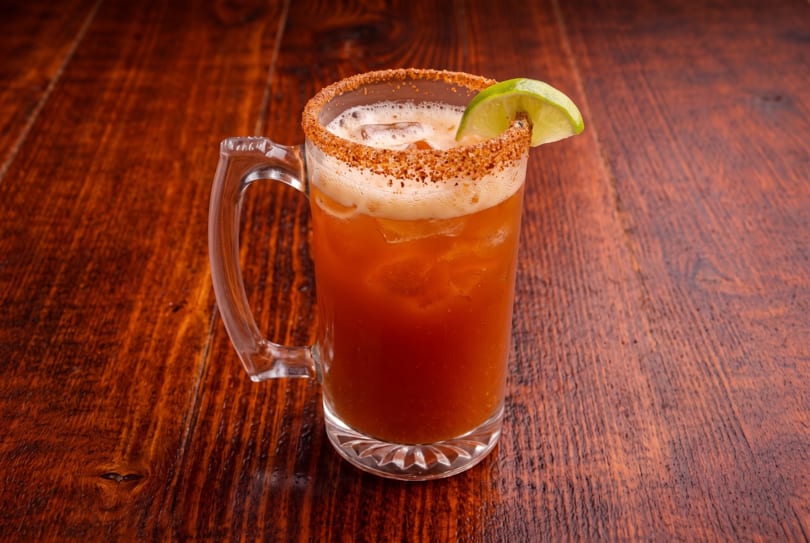Michelada in a glass mug with a spiced rim and lime wedge on a dark wooden table.