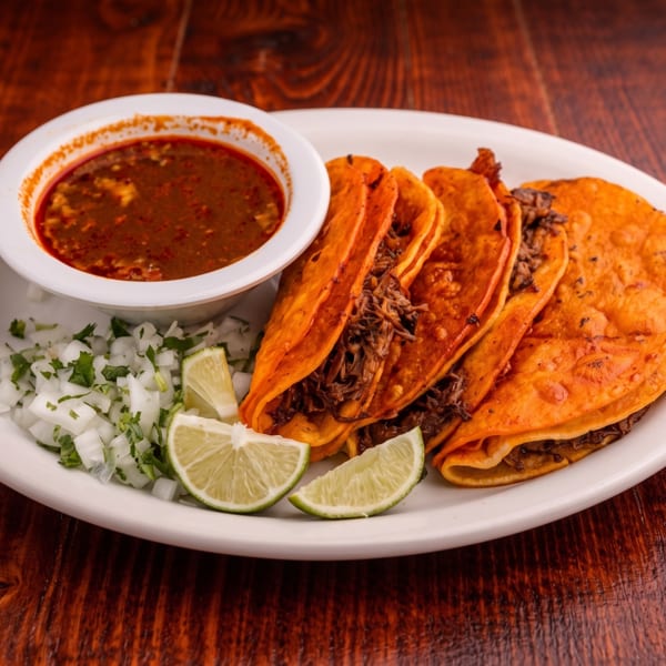 Four birria tacos with a side of consommé, diced onions, and lime on a white plate.