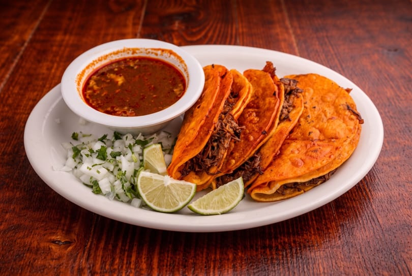 Four birria tacos with a side of consommé, diced onions, and lime on a white plate.