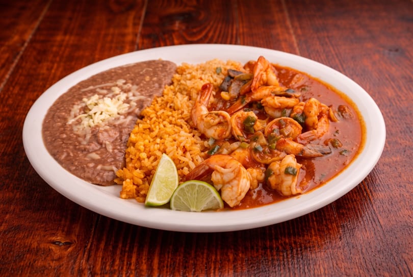 Shrimp in red sauce with rice, refried beans, and lime wedges on a white plate.