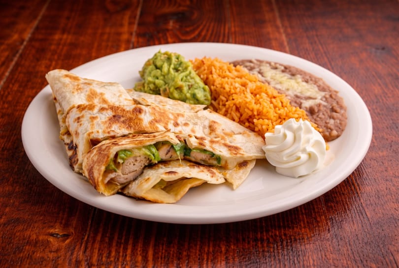 Chicken quesadillas with guacamole, Mexican rice, refried beans, and sour cream on a white plate.