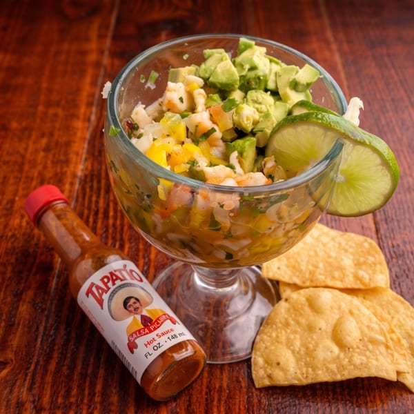 Ceviche in a glass with avocado, lime, Tapatio hot sauce, and tortilla chips on a wooden table.
