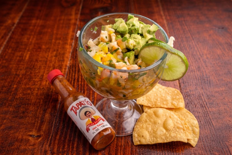 Ceviche in a glass with avocado, lime, Tapatio hot sauce, and tortilla chips on a wooden table.