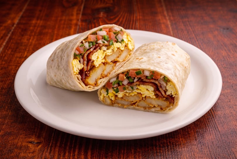 Breakfast burrito cut in half, showing eggs, bacon, hash browns, and pico de gallo on a white plate.