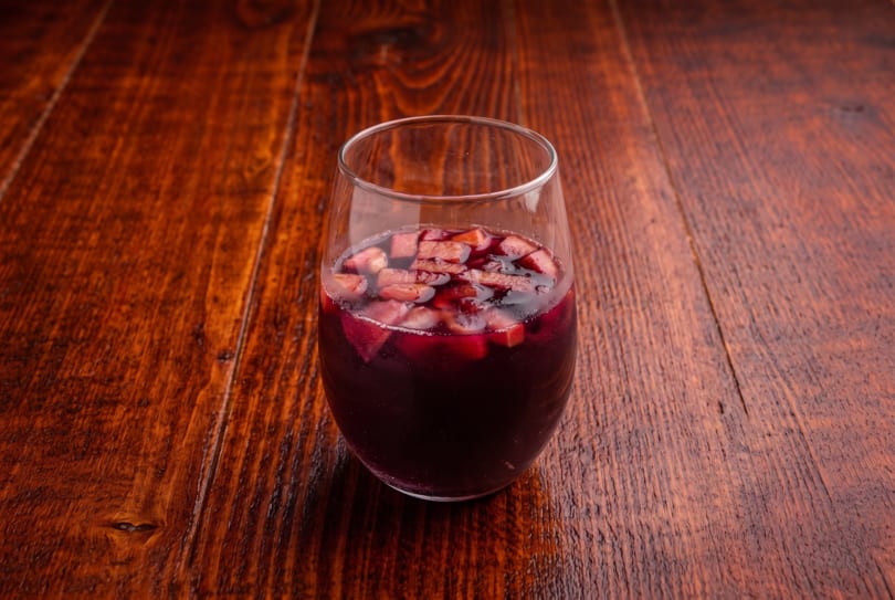 Glass of red sangria with diced fruit on a rustic wooden table.